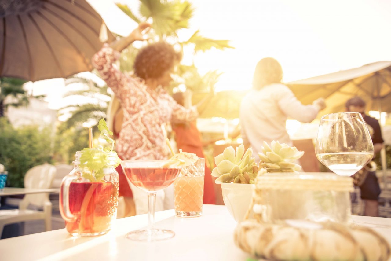 Tisch im Sonnenlicht mit verschiedenen Sommergetränken. Im Hintergrund tanzen Leute.